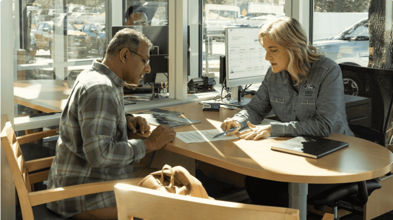 Two people reviewing documents together at a table inside a dealership or office setting.